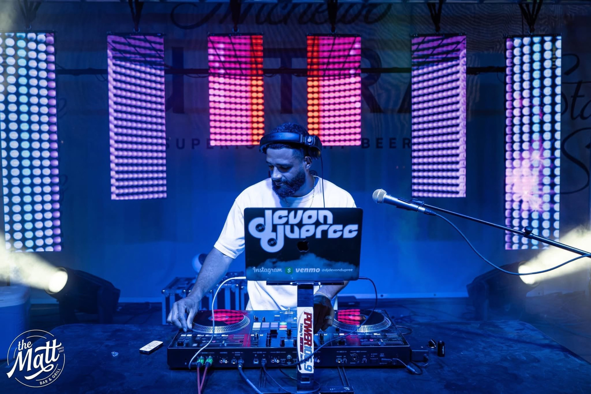 Devon Dupree mixing behind turntables under stage lighting at a nightlife venue